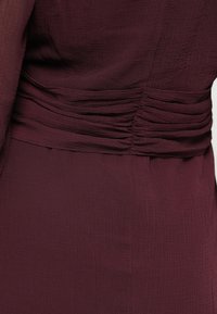 Μπορντό φόρεμα με υφασμάτινη υφή, λεπτομέρεια συγκέντρωσης στη μέση και ρέουσες μανσέτες. Λείο, ελαφρύ υλικό με διακριτό σχέδιο.