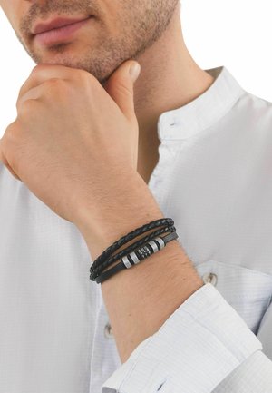 Mann in weißem Hemd stützt das Kinn auf die Hand und zeigt schwarze, geflochtene und Lederarmbänder mit Metallakzenten am Handgelenk.