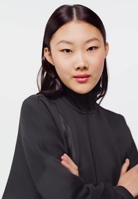 Mladi žena s ravnom tamnom kosom nosi crnu jaknu na patentni zatvarač, prekriženih ruku, neutralnog izraza lica, na jednostavnoj svijetloj pozadini.