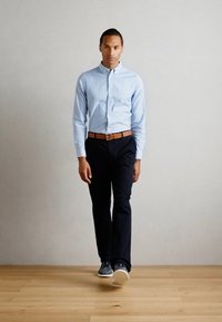 Helles blaues, knopfbares Hemd, schwarze Hose, brauer Gürtel und graue Sneaker. Das Modell steht vor einem schlichten hellen Hintergrund auf Holzboden.