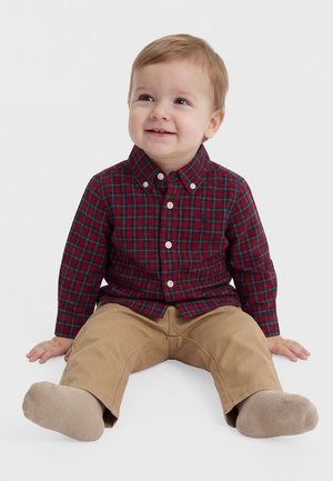Un tout-petit souriant est assis sur un fond blanc. Il porte une chemise à carreaux rouges et bleu marine, un pantalon beige et des chaussures beiges.