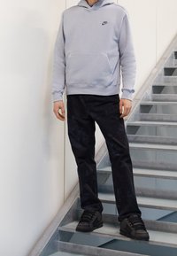 Světlý šedý mikina Nike s přední kapsou a černým logem, v kombinaci s tmavými vzorovanými kalhotami a černými sportovními botami, stojící na schodech.