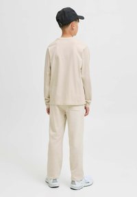 Junge trägt ein cremefarbenes Langarmshirt und eine Hose, weiße Sneakers und eine schwarze Baseballmütze, die von einem einfachen weißen Hintergrund abgewandt ist.