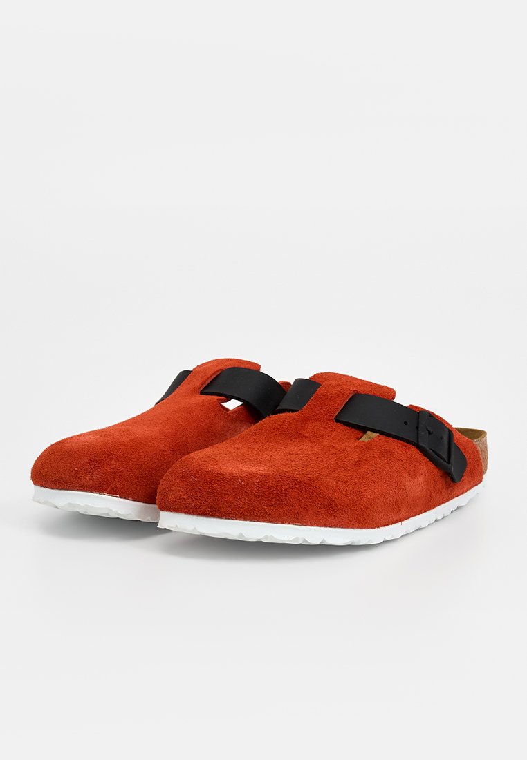 Rote Wildleder-Clogs mit einem schwarzen Riemen, runden Zehen und einer weißen, strukturierten Gummisohle für besseren Halt.