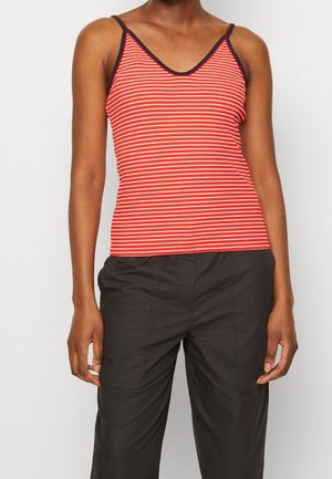Persona vestida con una camiseta sin mangas a rayas rojas y blancas con ribete negro y pantalones negros holgados, de pie frente a un fondo liso.