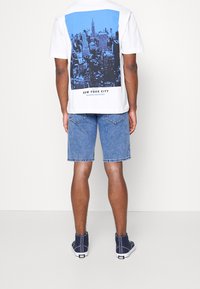 Camiseta blanca de manga corta con un gráfico de la silueta de Nueva York en azul en la parte posterior, combinada con pantalones cortos de mezclilla azules y zapatillas altas navy.