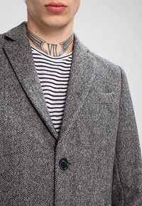 Blazer grigio a motivo houndstooth con rever a scollo, chiusura con un bottone e tasca decorativa. Camicia interna a righe con linee orizzontali bianche e nere.