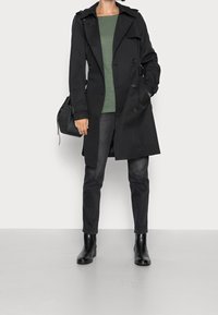 Zwarte trenchcoat met dubbele knopen, riem en schouderflappen. Gecombineerd met een groene shirt, donkere spijkerbroek en zwarte enkellaarsjes.