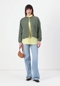 Grøn quilted jakke med frontknaplukning og sidelommer, båret over en gul skjorte, parret med flagrende blå jeans og sorte flade sko.