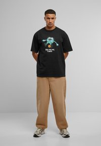Camiseta de algodón negra con un diseño de rana de dibujos animados. El texto dice "Llévame afuera, entrenador." Combinada con pantalones beige de corte holgado y zapatillas.