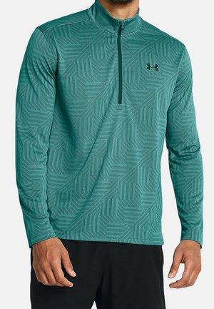 Man draagt een teal lange mouw sportshirt met een kwart-ritssluiting, geometrisch patroon en zwart logo op de borst, gecombineerd met een zwarte broek.