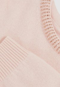 Hellrosa Pullover mit gerippten Bündchen und Ausschnitt, verziert mit einer Reihe von roségoldenen Perlen um den Kragen. Weiche Stricktextur.