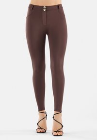 Leggings marroni a vita alta con una texture liscia, caratterizzati da una chiusura frontale con bottoni e tasche laterali. Abbinati a tacchi neri con cinturini.