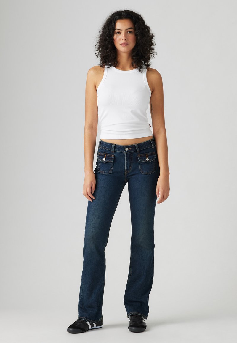 Schlagjeans aus dunkelblauem Denim mit Fronttaschen und metallischem Knopfverschluss. Kombiniert mit einem weißen gerippten Tanktop und schwarzen Schuhen.