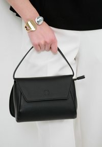Borsa a mano in pelle nera con manico superiore, texture liscia, chiusura a pattina e logo in rilievo sulla parte anteriore. Accompagnata da un bracciale metallico.