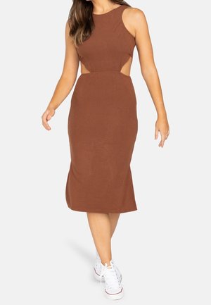 Femme marchant portant une robe midi marron sans manches avec des découpes latérales et des baskets blanches à lacets sur un fond blanc.