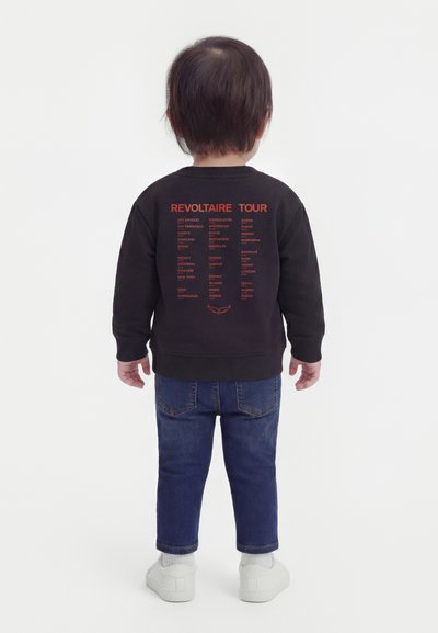 Παιδί που στέκεται με την πλάτη στραμμένη προς τον θεατή, φοράει μαύρο φούτερ με κόκκινο κείμενο "REVOLTAIRE TOUR" και ονόματα πόλεων, μπλε τζιν και λευκά αθλητικά παπούτσια.