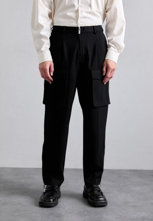 Μαύρο cargo παντελόνι με μπροστινές τσέπες, φερμουάρ και χαλαρή γραμμή. Συνδυάζεται με ένα ανοιχτόχρωμο κουμπωτό πουκάμισο και μαύρα παπούτσια.