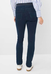 Jeans blu scuro elasticizzati con design a cinque tasche, vita alta e vestibilità slim, abbinati a scarpe bianche e argentate su uno sfondo minimale.