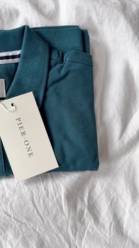 Polo di colore teal piegato ordinatamente, realizzato in tessuto testurizzato, con un'etichetta con la scritta "PIER ONE" attaccata, posizionato su una superficie bianca sgualcita.