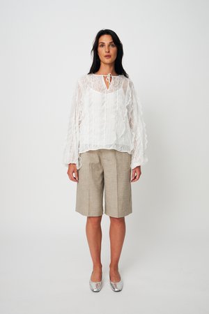 Γυναίκα που φοράει λευκή δαντελένια μπλούζα με μακριά μανίκια, μπεζ σορτς μέχρι το γόνατο και ασημένια φλατ παπούτσια, στέκεται μπροστά σε απλό φωτεινό φόντο.