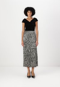 Schwarzes Samtop-Top mit Twist-Detail, kombiniert mit einem langen weißen Rock mit einem schwarzen abstrakten Muster. Das Modell trägt schwarze Absätze.
