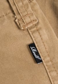 Close-up of beige fabric seam with belt loop and small black clothing label attached to the stitching.