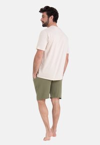 Chemise beige à manches courtes associée à un short texturé vert olive. Le modèle se tient pieds nus, les mains dans les poches, vu de dos.