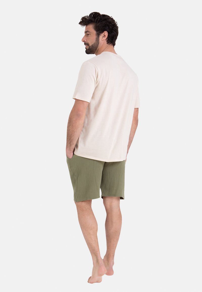 Chemise beige à manches courtes associée à un short texturé vert olive. Le modèle se tient pieds nus, les mains dans les poches, vu de dos.