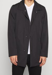 Czarny płaszcz przeciwdeszczowy z usztywnionym kołnierzem, zapięciem na guziki, bocznymi kieszeniami i gładką, matową fakturą. Noszony na białej koszulce.