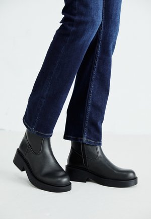 Personne portant un jean bleu foncé et des bottines en cuir noir à semelles épaisses, debout sur un sol blanc avec un fond blanc uni.