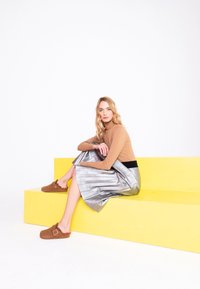 Tan turtleneck, shiny silver pleated skirt with black waistband, and brown suede slip-on shoes with decorative buckle, seated on yellow steps.
