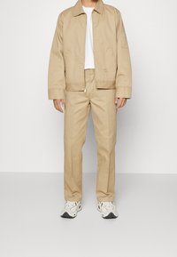 Beige bomullsjacka med krage, dragkedja fram och sidofickor, ihop med matchande byxor. Lättviktig tyg, enkel design och avslappnad passform.