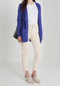 Blå stickad cardigan med öppen front och fickor, kombinerad med vit t-shirt och beige jeans med fransar. Grå muleskor. Svart handväska.