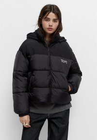Giubbotto puffer nero con cappuccio, caratterizzato da una zip frontale, polsini elasticizzati e un piccolo logo sul petto. Design leggero e trapuntato.
