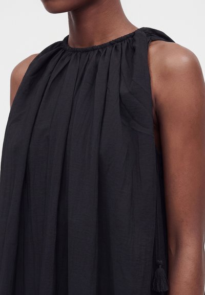 Femme portant une robe noire sans manches avec un col froncé et un lien à pompons sur l'épaule, montrant le haut de la poitrine et le bras.