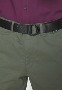 Pantaloni verde oliva con una texture liscia, dotati di una cintura in tessuto nera e una fibbia. Cuciture dettagliate visibili lungo le cuciture.