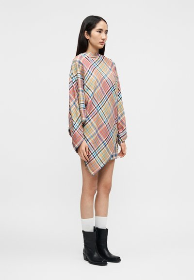 Γυναίκα που στέκεται, φορώντας πολύχρωμο ασύμμετρο καρό φόρεμα, λευκές κάλτσες και μαύρα μπότες μέχρι τη μέση της γάμπας, μπροστά σε απλό ανοιχτό γκρι φόντο.