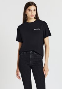 Giovane donna con lunghi capelli castani che indossa una maglietta nera di "Sex and the City" e jeans scuri su uno sfondo bianco semplice.