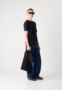 Black t-shirt, oversized blue denim pants, black sunglasses, holding a black hoodie, casual footwear, smooth fabric, relaxed fit.