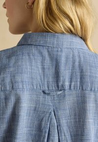 Blauw-wit geruit katoenen shirt, met een kraag, een rugplooi en een lusdetail onder de kraag. Glad van structuur.