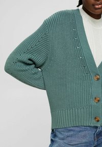 Zielony dziergany kardigan z głębokim dekoltem w serek, z zapięciem na guziki i ściągaczami na mankietach. Posiada teksturowany materiał oraz małe otworki wzdłuż rękawów.