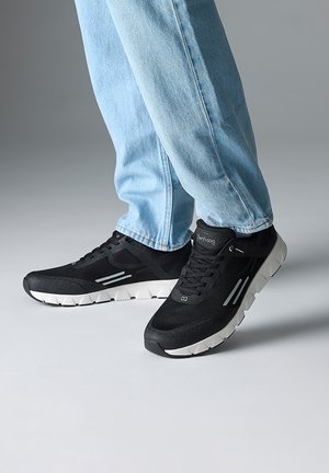 Person trägt hellblaue Jeans und schwarze Sportschuhe mit weißen Sohlen und steht auf einer grauen Oberfläche.