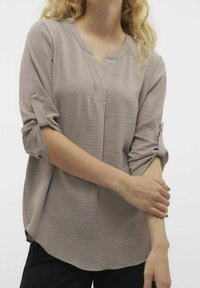 Blusa di un taupe chiaro con finitura testurizzata, scollatura a V e maniche a tre quarti, con bottoni per arrotolare le maniche e vestibilità ampia.