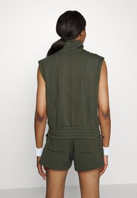 Mujer con cabello oscuro en una coleta, vistiendo una camiseta sin mangas de color verde oscuro y pantalones cortos a juego, de espaldas, con pulseras blancas en ambas muñecas.