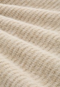 Beige gestrickter Stoff mit strukturiertem Rippenmuster und einer weichen, flauschigen Textur, die ein warmes, natürliches Erscheinungsbild mit subtilen Tonvariationen schafft.