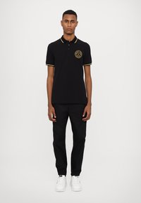 Cămașă polo neagră cu manșete și guler accent galben, având un logo circular pe piept. Purtați cu pantaloni negri și adidași albi.