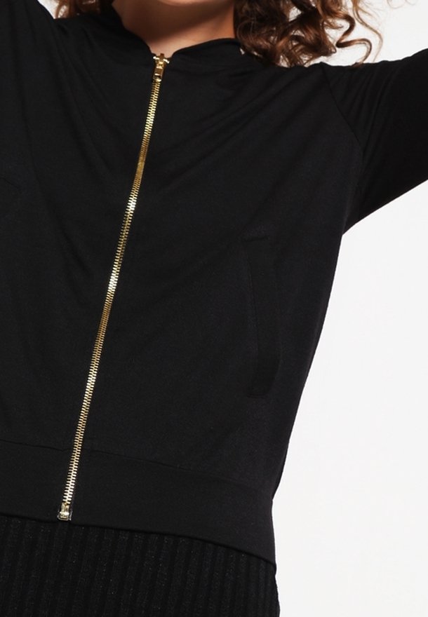 Musta vetoketjullinen takki, joka on valmistettu sileästä kankaasta. Siinä on kultainen vetoketju ja kaksi etutaskua, sekä muotoiltu vyötärö ja pitkät hihat.