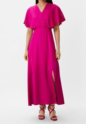 Femme portant une longue robe rose vif avec des manches volantées, un col en V, une fente latérale et des sandales à talons hauts assorties avec des lanières.