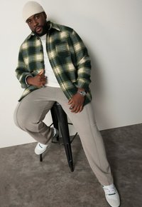 Camicia flanella a quadri verdi e crema, pantaloni grigi, t-shirt bianca, sneaker bianche con dettagli verdi, seduto su uno sgabello nero.